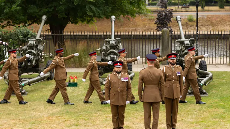 A group of uniformed soldiers stands and walks in formation on a grassy area in front of ceremonial cannons.