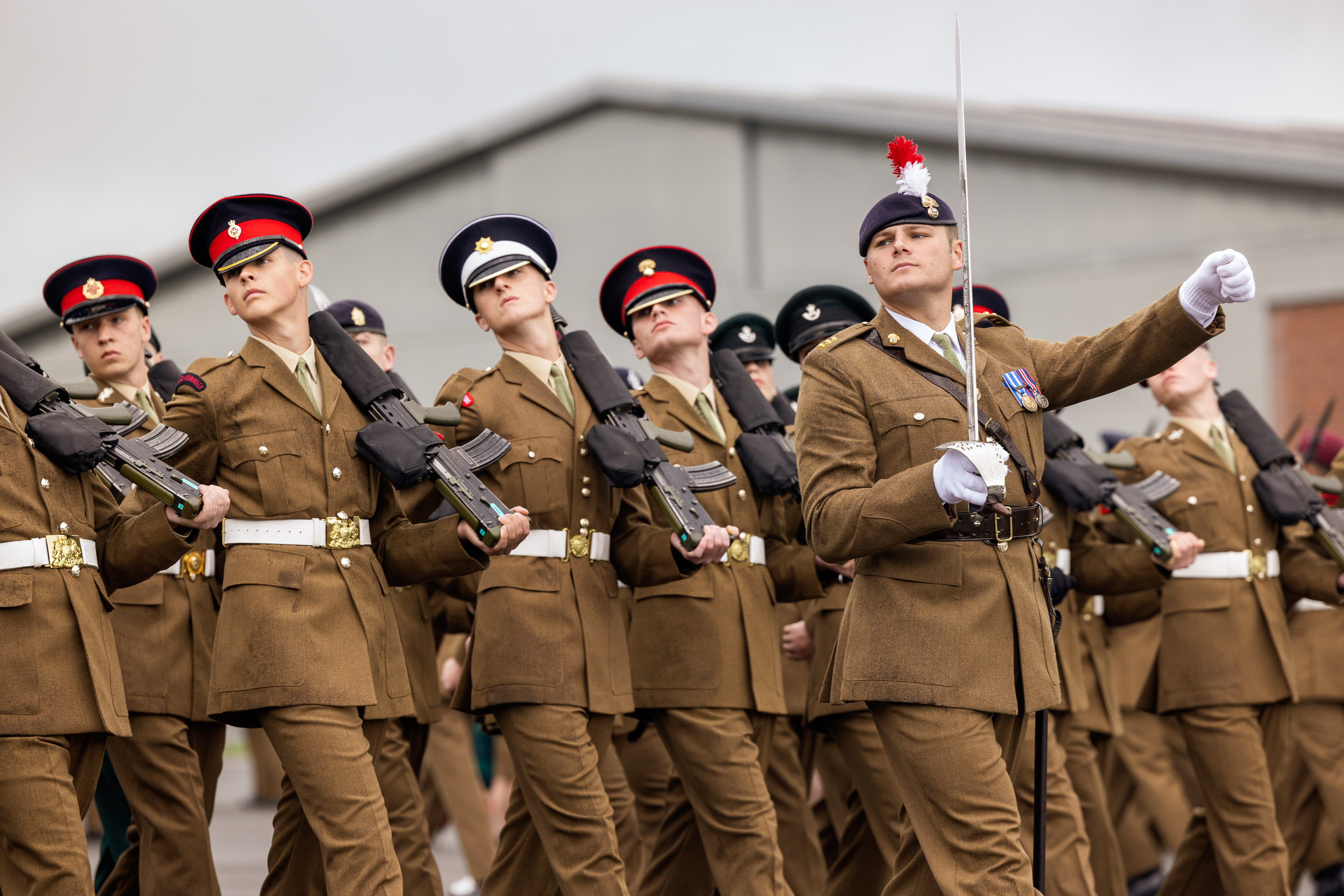 A row of soldiers marching in line wearing brown uniforms and holding a rifle. They are marching forward with their heads turned looking to their right.