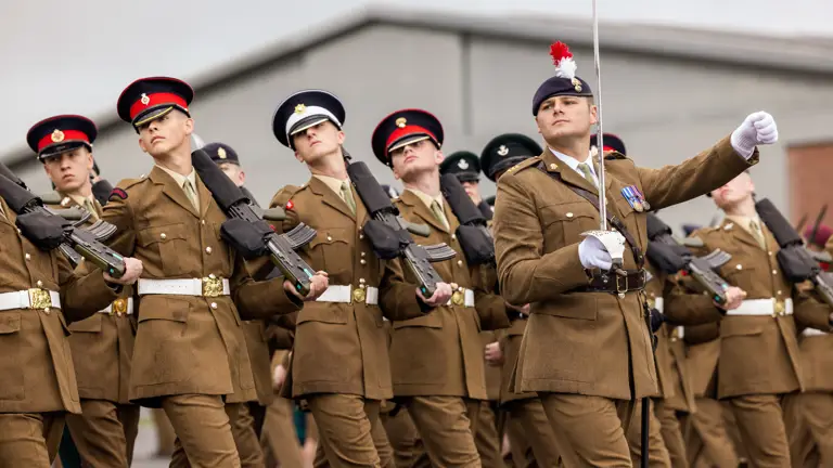 A row of soldiers marching in line wearing brown uniforms and holding a rifle. They are marching forward with their heads turned looking to their right.