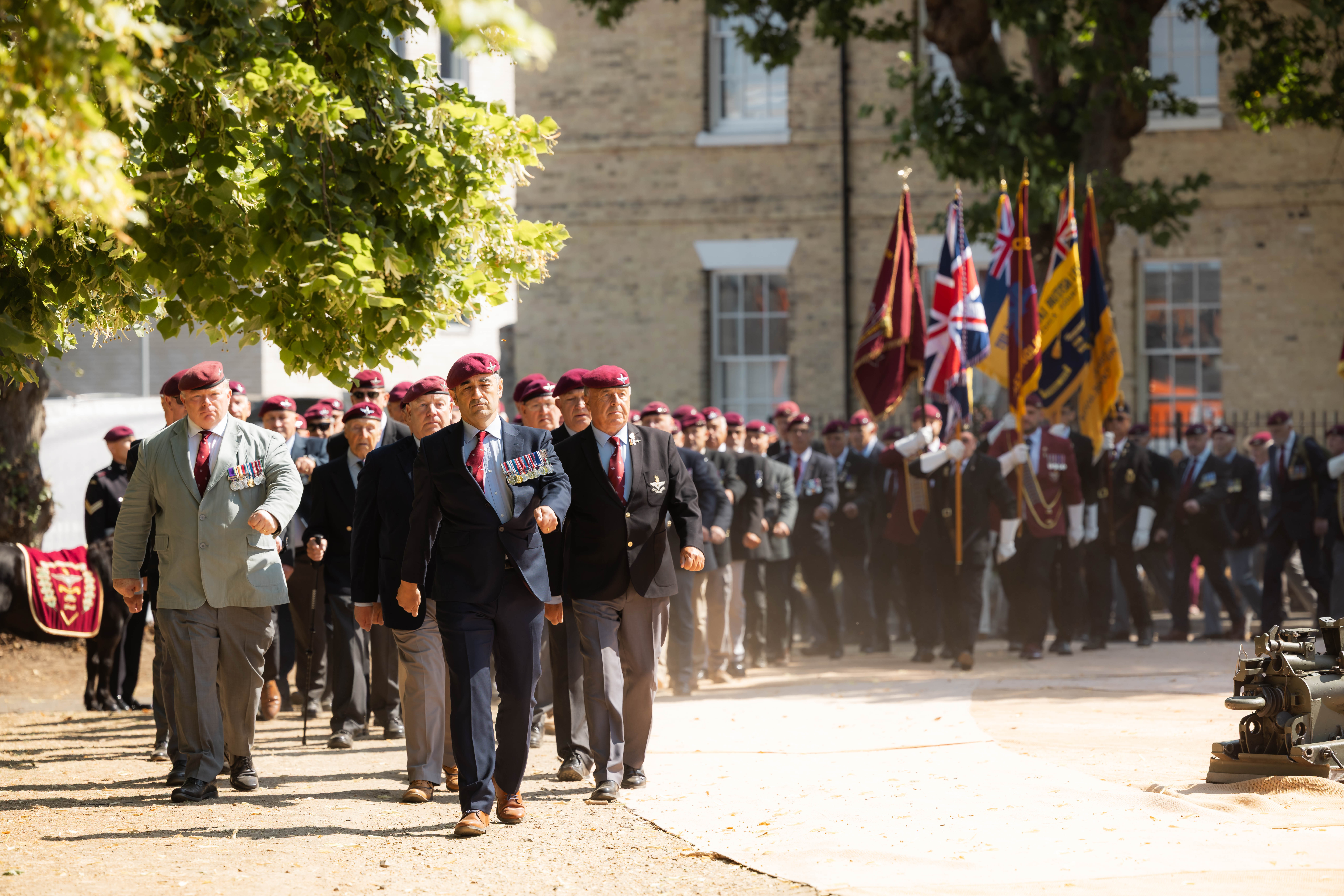 A group of people in uniform, wearing maroon berets and medals, are marching in formation. Behind them, others carry a line of flags, including the Union Jack. They are walking on a path with trees and a brick building in the background.