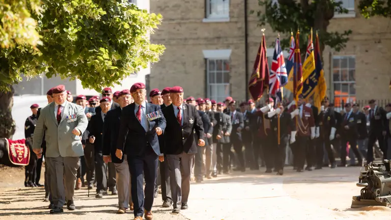 A group of people in uniform, wearing maroon berets and medals, are marching in formation. Behind them, others carry a line of flags, including the Union Jack. They are walking on a path with trees and a brick building in the background.