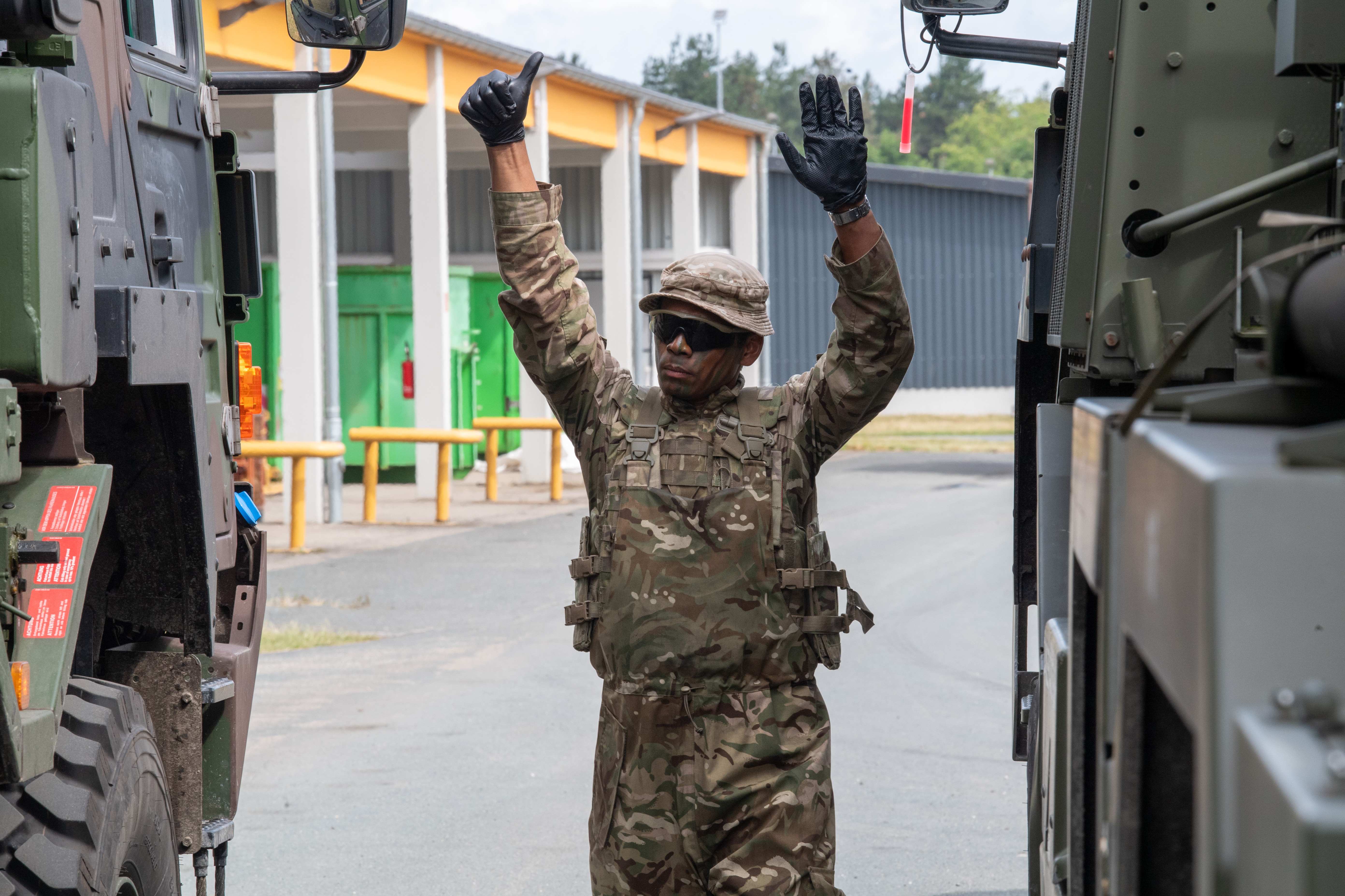 Soldier in camouflage uniform and tactical vest raising both hands between two military vehicles in an outdoor setting.