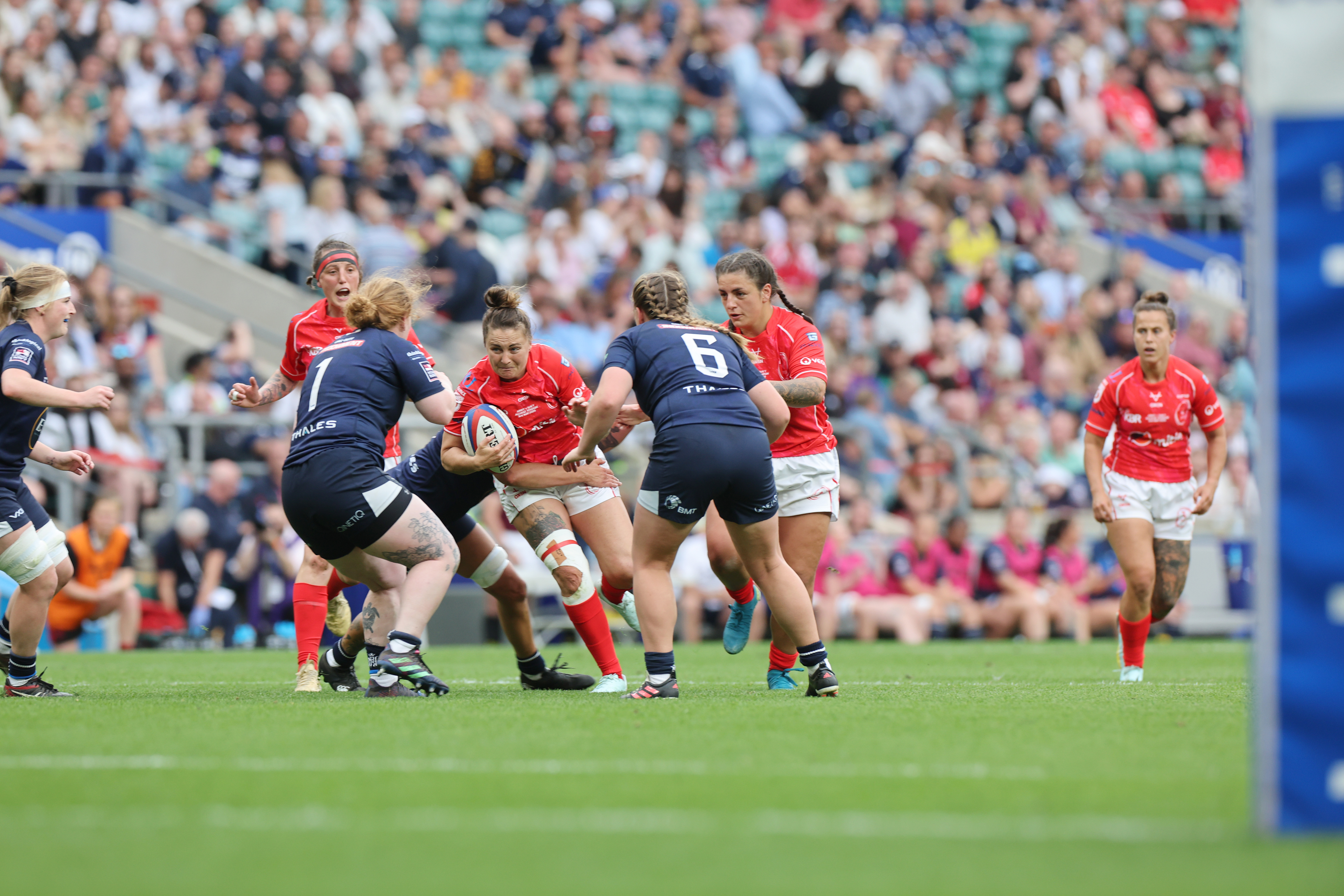 Players in red and blue rugby kits engage in a tense match on a green field. A full stadium of spectators watches intently in the background.