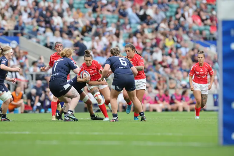 Players in red and blue rugby kits engage in a tense match on a green field. A full stadium of spectators watches intently in the background.