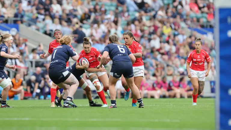 Players in red and blue rugby kits engage in a tense match on a green field. A full stadium of spectators watches intently in the background.