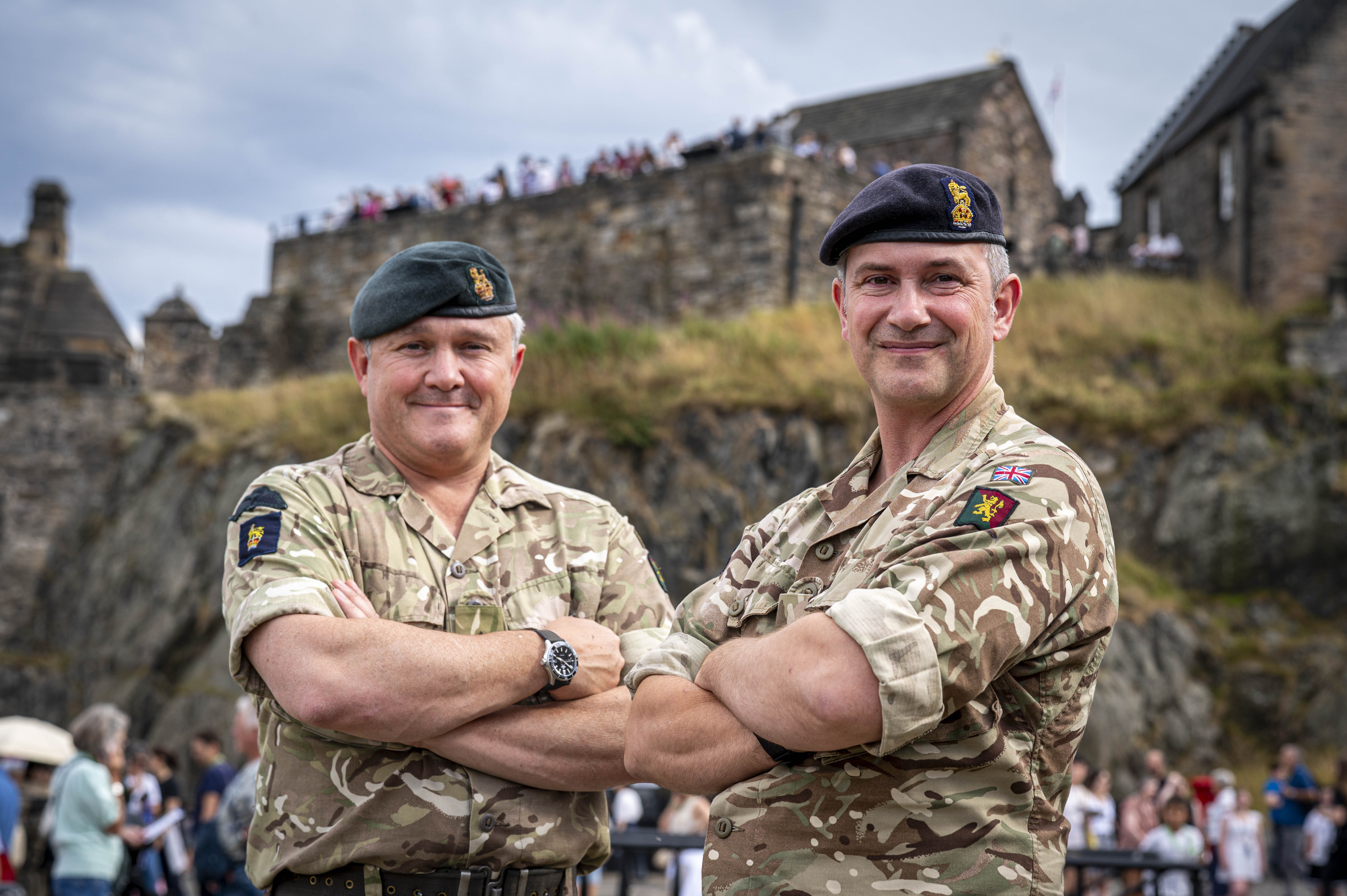 Two individuals in military uniform, each wearing a beret with insignia, standing confidently with arms crossed. They are outdoors with an architectural structure visible in the background.