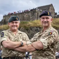 Two individuals in military uniform, each wearing a beret with insignia, standing confidently with arms crossed. They are outdoors with an architectural structure visible in the background.