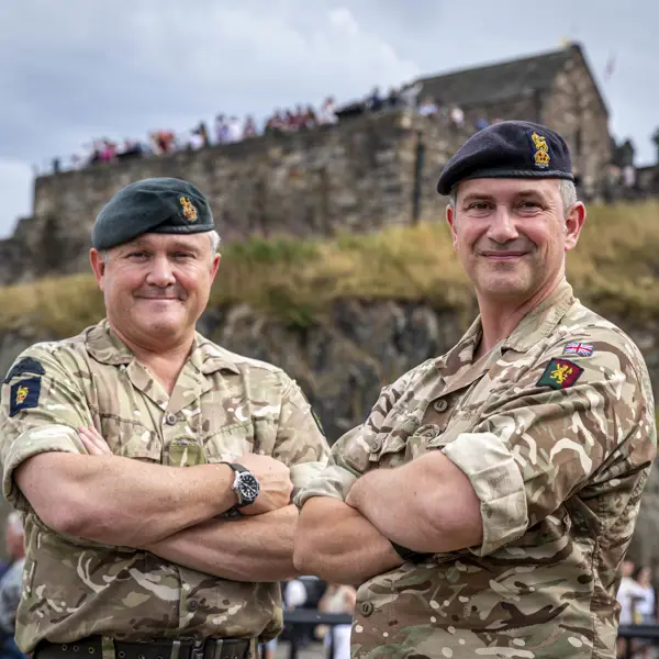 Two individuals in military uniform, each wearing a beret with insignia, standing confidently with arms crossed. They are outdoors with an architectural structure visible in the background.