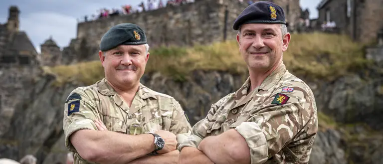 Two individuals in military uniform, each wearing a beret with insignia, standing confidently with arms crossed. They are outdoors with an architectural structure visible in the background.