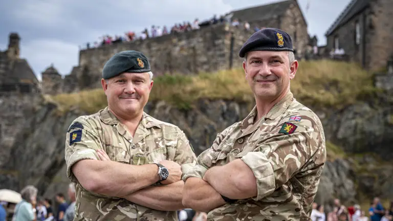 Two individuals in military uniform, each wearing a beret with insignia, standing confidently with arms crossed. They are outdoors with an architectural structure visible in the background.