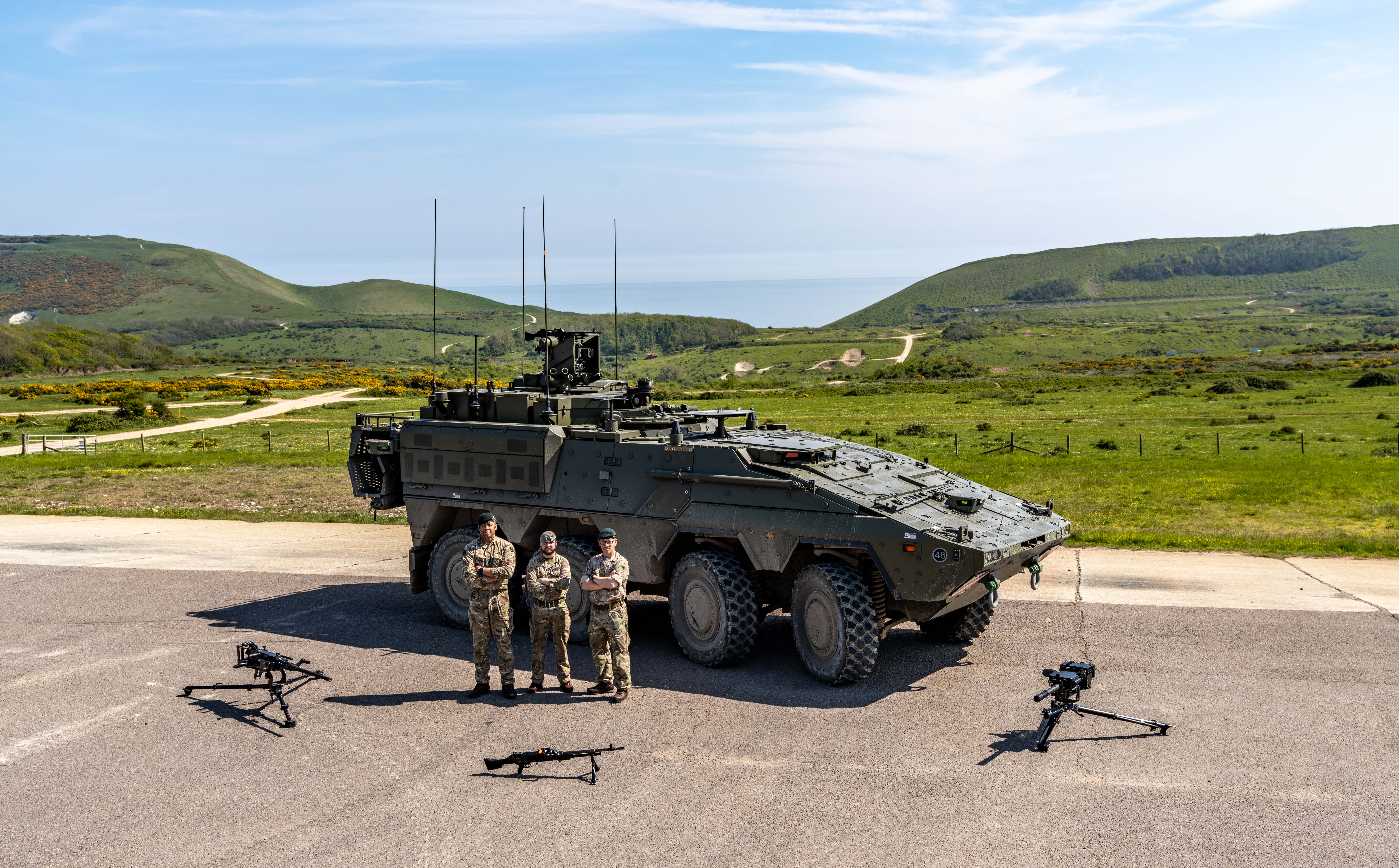 A group of military personnel stands in front of an armoured vehicle, a Boxer, on a paved area. 