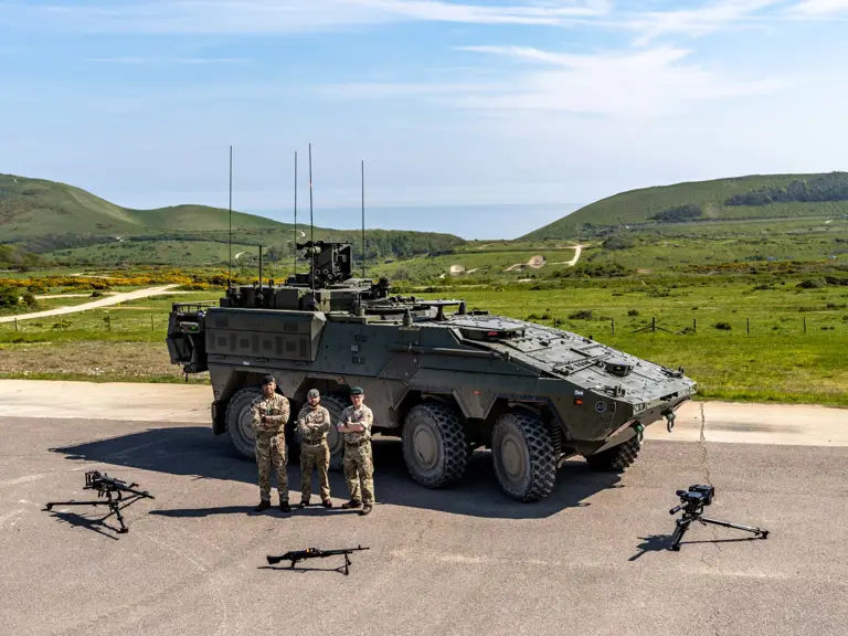A group of military personnel stands in front of an armoured vehicle, a Boxer, on a paved area.