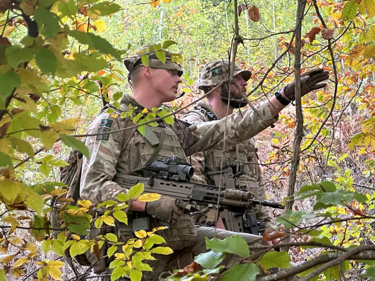 Two soldiers stand behind some branches. One of the soldiers is pointing.