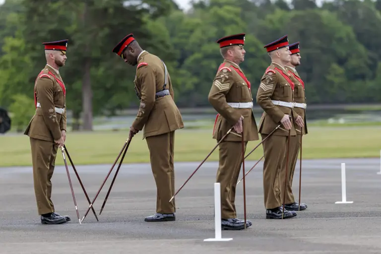 Soldiers in formal brown uniforms and red hats stand in formation on a paved area, practicing drill maneuvers with rifles. They are positioned in front of a backdrop of trees and greenery.