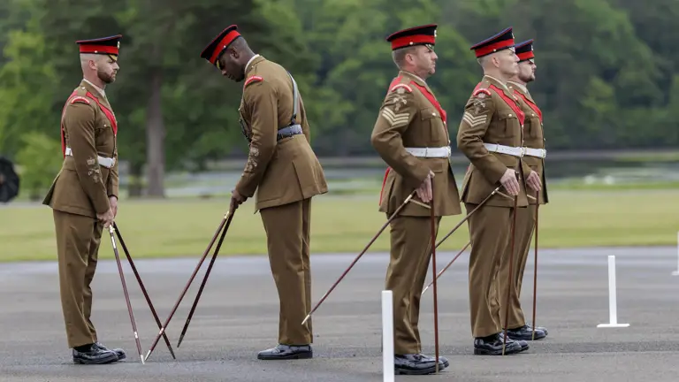 Soldiers in formal brown uniforms and red hats stand in formation on a paved area, practicing drill maneuvers with rifles. They are positioned in front of a backdrop of trees and greenery.