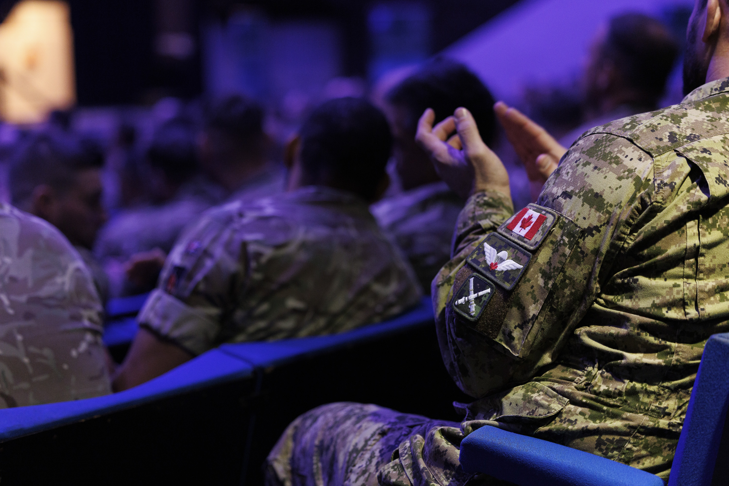 A group of soldiers in camouflage uniforms sit in auditorium chairs. One soldier's sleeve displays a Canadian flag patch, suggesting a formal or official event.