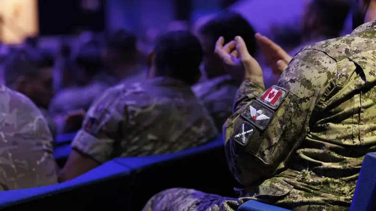 A group of soldiers in camouflage uniforms sit in auditorium chairs. One soldier's sleeve displays a Canadian flag patch, suggesting a formal or official event.