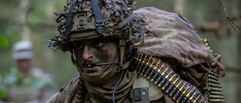 Soldier in camouflage gear with a helmet covered in foliage and multiple belts of ammunition across the chest.