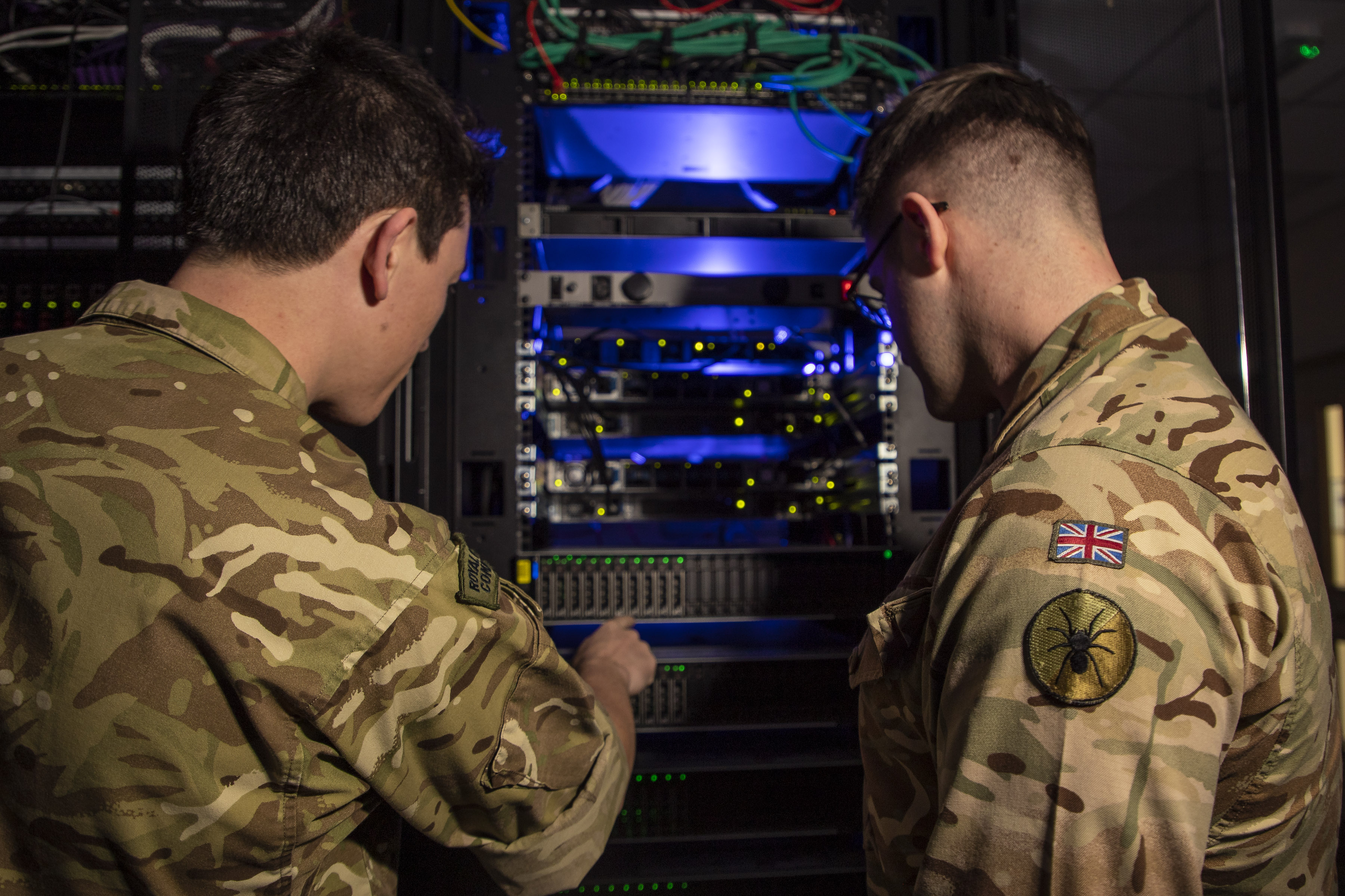 Two soldiers in camouflage uniforms inspect a rack of illuminated network servers in a dimly lit room.