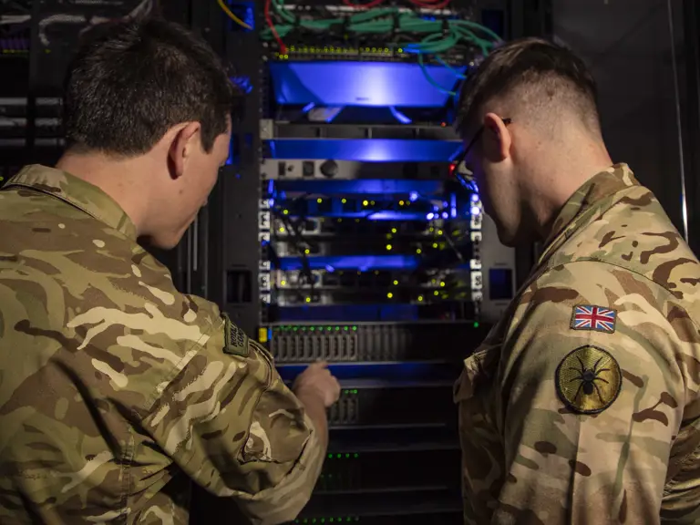 Two soldiers in camouflage uniforms inspect a rack of illuminated network servers in a dimly lit room.