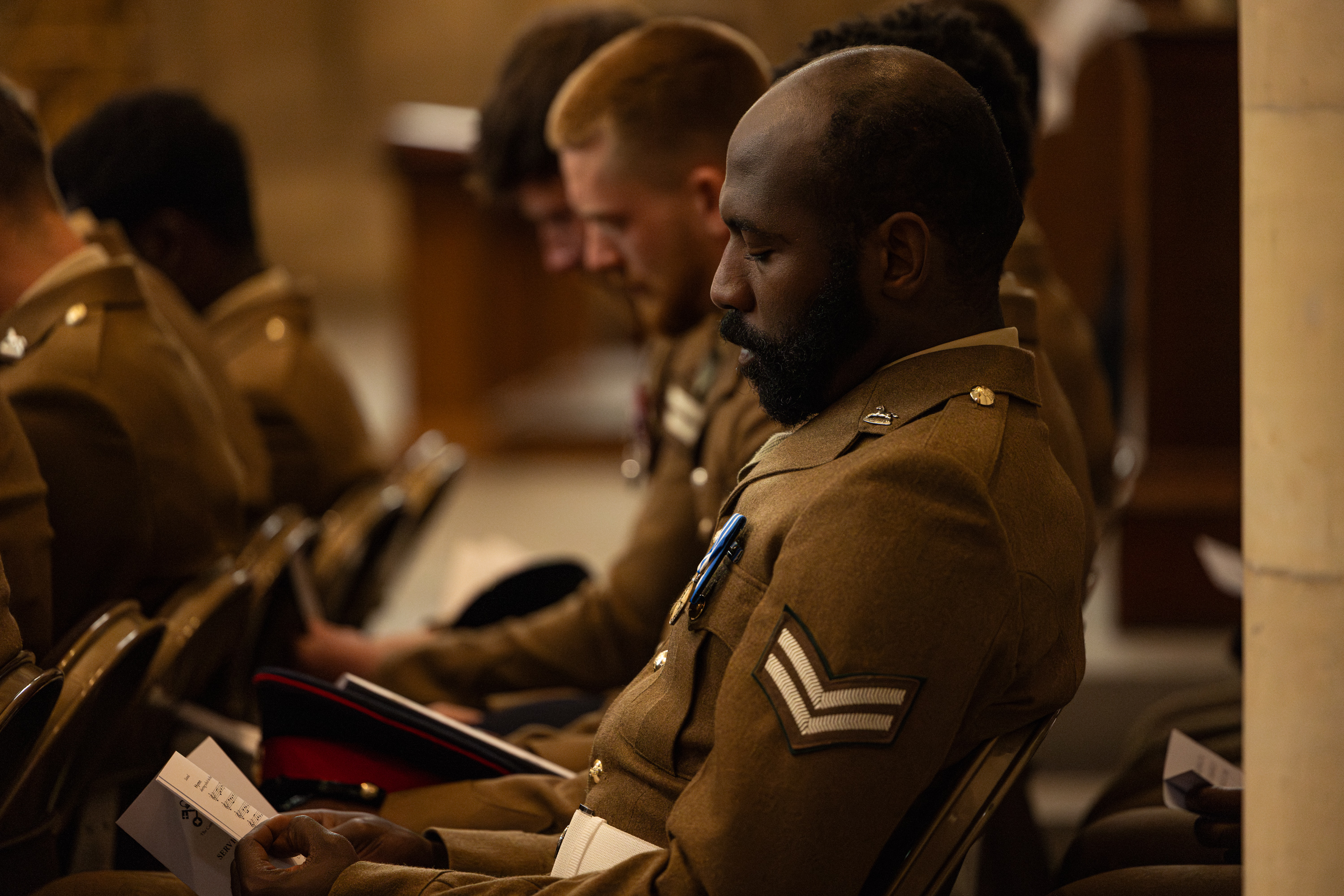 A group of individuals in military uniforms are seated indoors, holding programs, and appear focused on an event or ceremony. The setting suggests a formal or commemorative occasion.