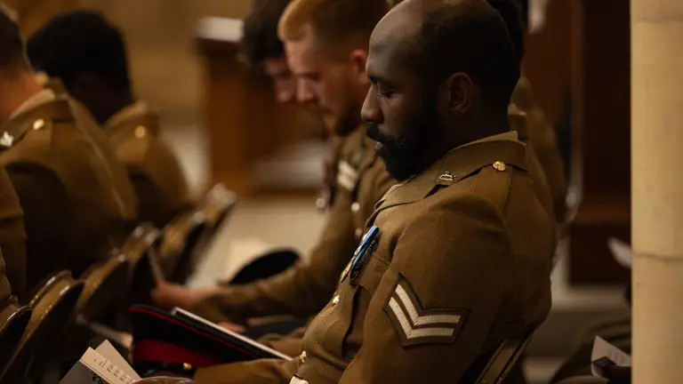 A group of individuals in military uniforms are seated indoors, holding programs, and appear focused on an event or ceremony. The setting suggests a formal or commemorative occasion.