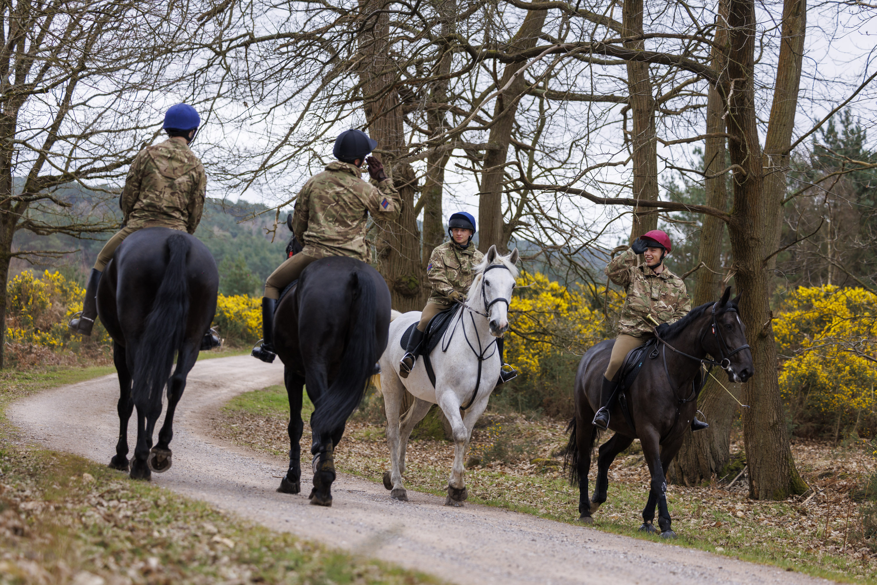 A group of people in camouflage uniforms are riding horses along a forest path. The horses vary in colour.