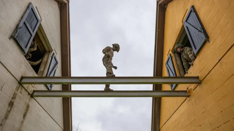Soldier in camouflage uniform and helmet seen balancing across steel girders from house to house.