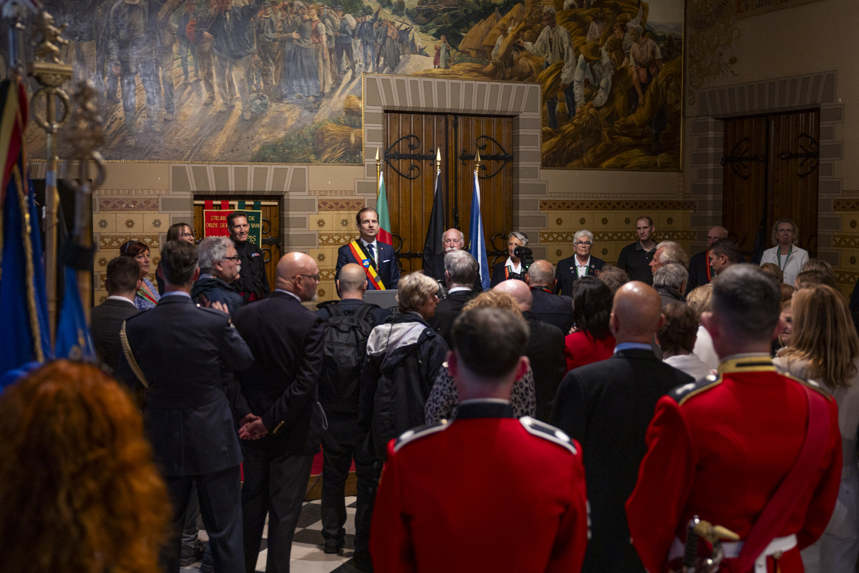 Crowd gathered in a historic hall with ornate murals and flags, listening to a speaker at a podium.