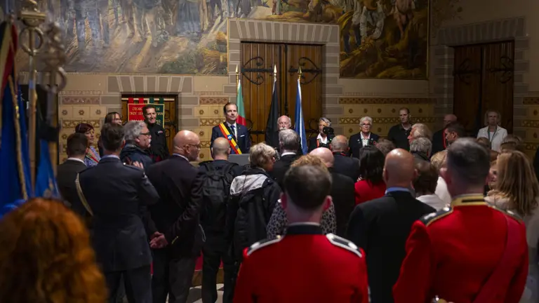 Crowd gathered in a historic hall with ornate murals and flags, listening to a speaker at a podium.