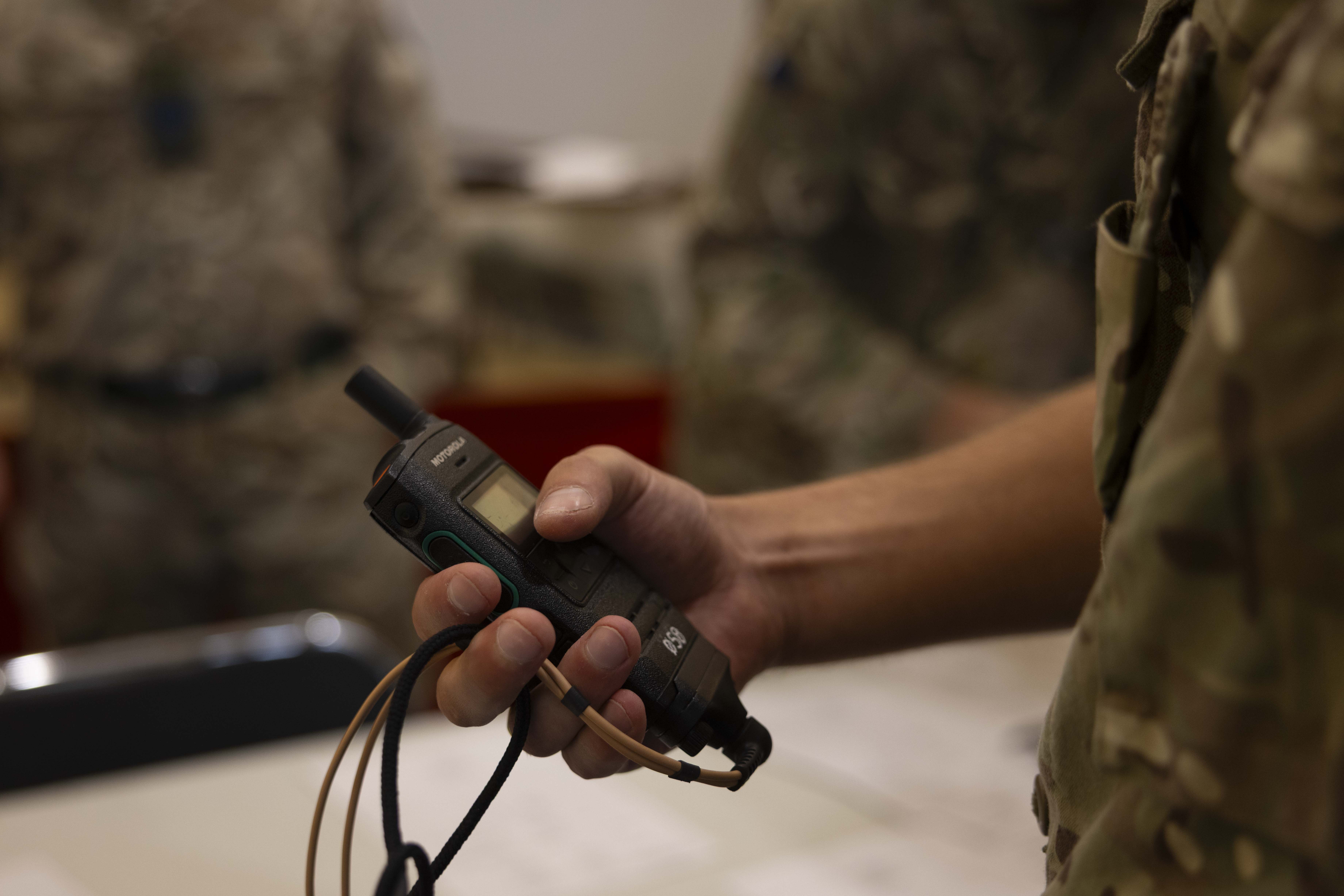 A soldier in camouflage holds a military radio device in hand, suggesting communication readiness. Background shows other soldiers, slightly out of focus.
