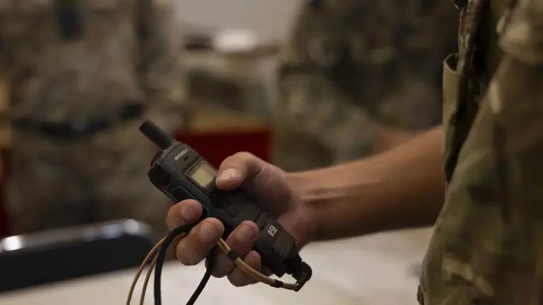 A soldier in camouflage holds a military radio device in hand, suggesting communication readiness. Background shows other soldiers, slightly out of focus.