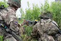 Two soldiers in camouflage gear crouch in tall grass, one feeding ammunition belt into a machine gun held by the other.