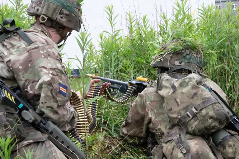 Two soldiers in camouflage gear crouch in tall grass, one feeding ammunition belt into a machine gun held by the other.
