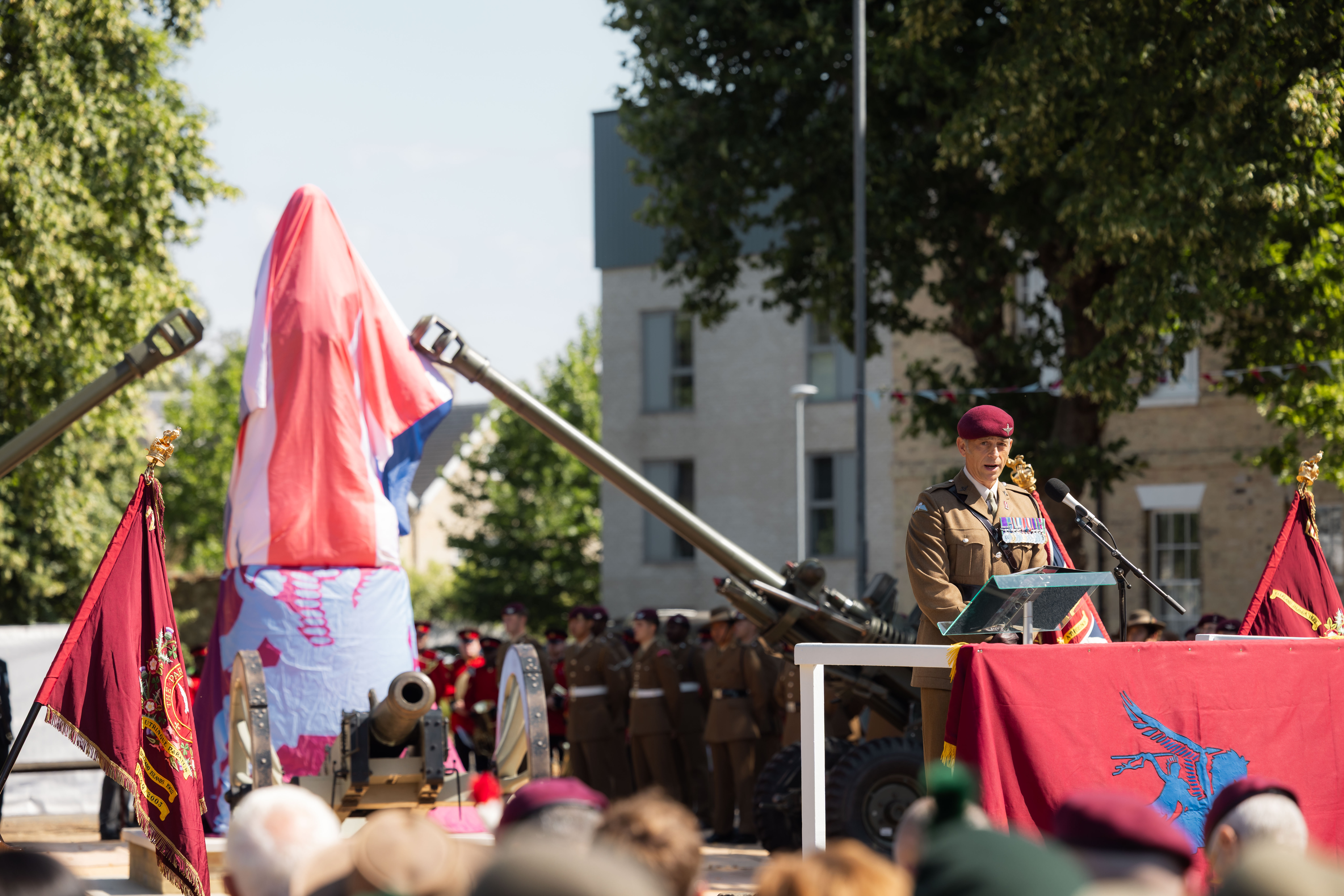 Man in brown uniform and a red beret stood behind a red banner talking to a crowd, the covered statue behind him between two big guns.