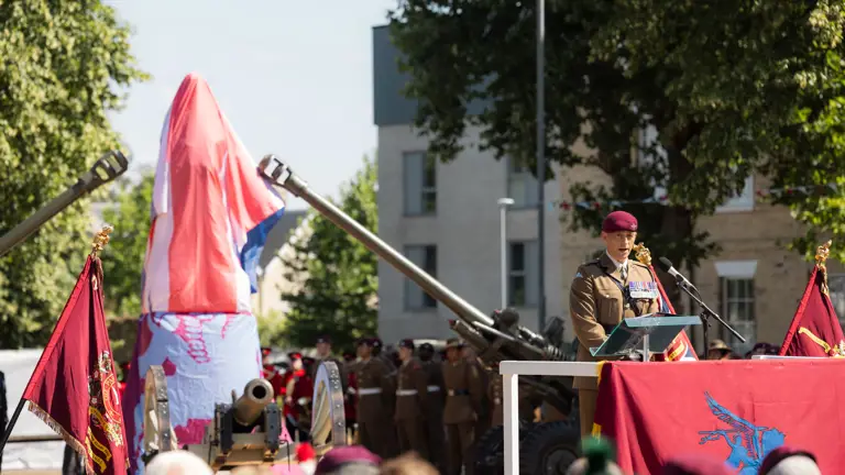 Man in brown uniform and a red beret stood behind a red banner talking to a crowd, the covered statue behind him between two big guns.