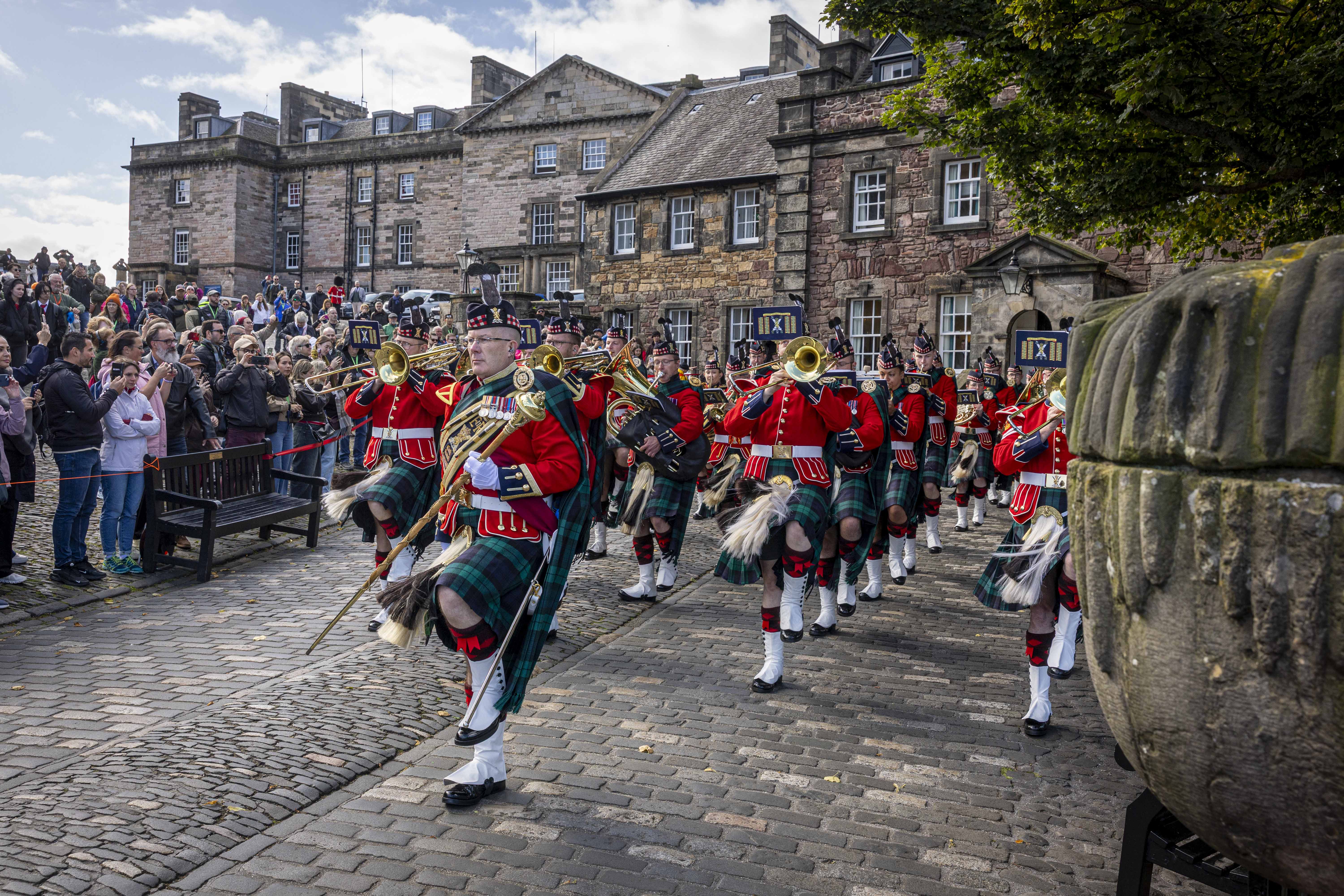 A marching band in traditional Scottish attire, including kilts and red jackets, performs with brass instruments in a cobbled courtyard. A large crowd watches, and historic stone buildings are visible in the background.