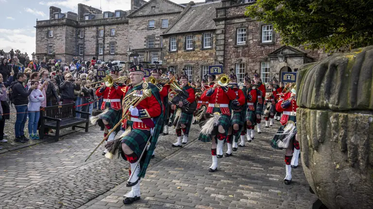 A marching band in traditional Scottish attire, including kilts and red jackets, performs with brass instruments in a cobbled courtyard. A large crowd watches, and historic stone buildings are visible in the background.