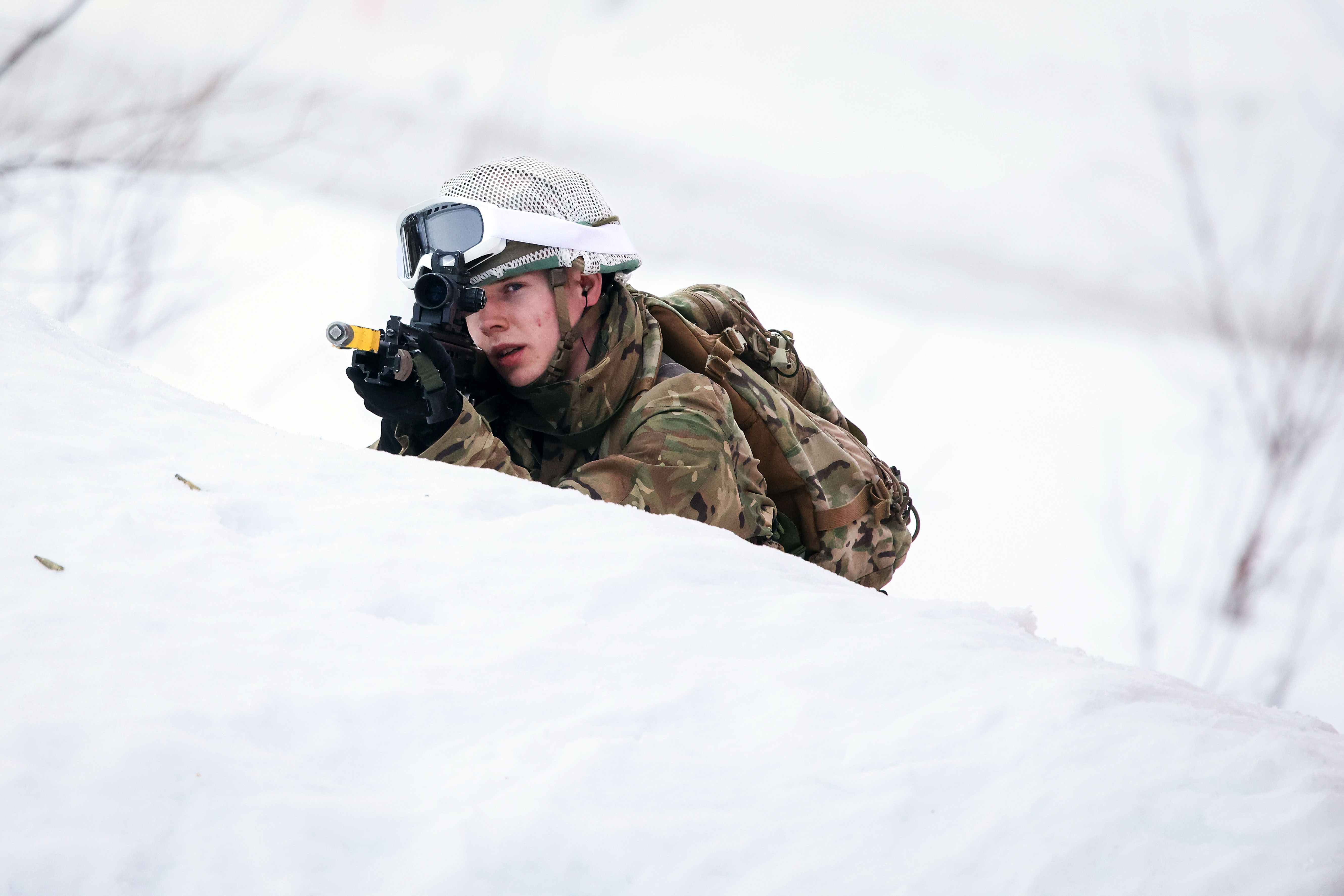 Soldier in camouflage gear aiming a rifle while lying in deep snow during winter training.