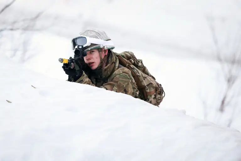 Soldier in camouflage gear aiming a rifle while lying in deep snow during winter training.