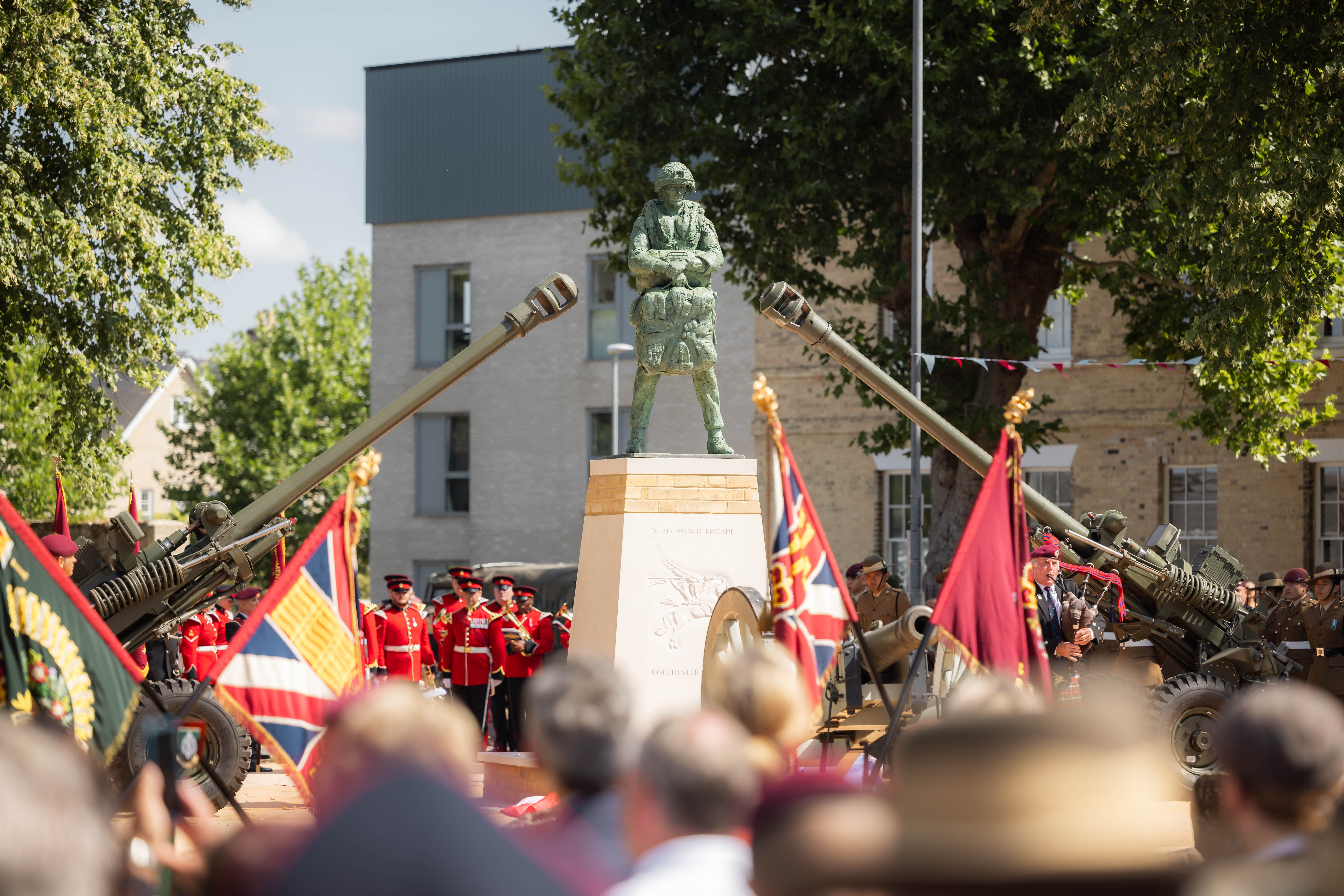 A ceremonial event with uniformed individuals standing in formation around a monument. The scene includes decorative flags and two artillery guns pointed upward. The setting is outdoors, surrounded by trees and buildings.