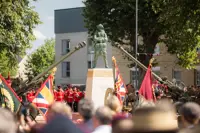 A ceremonial event with uniformed individuals standing in formation around a monument. The scene includes decorative flags and two artillery guns pointed upward. The setting is outdoors, surrounded by trees and buildings.
