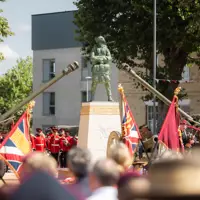 A ceremonial event with uniformed individuals standing in formation around a monument. The scene includes decorative flags and two artillery guns pointed upward. The setting is outdoors, surrounded by trees and buildings.