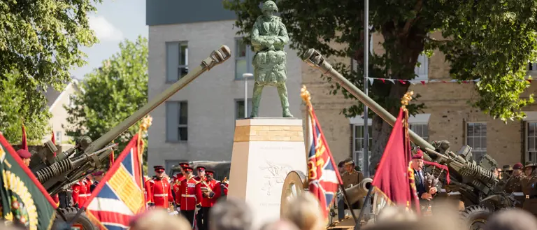 A ceremonial event with uniformed individuals standing in formation around a monument. The scene includes decorative flags and two artillery guns pointed upward. The setting is outdoors, surrounded by trees and buildings.