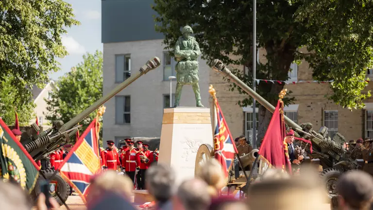 A ceremonial event with uniformed individuals standing in formation around a monument. The scene includes decorative flags and two artillery guns pointed upward. The setting is outdoors, surrounded by trees and buildings.