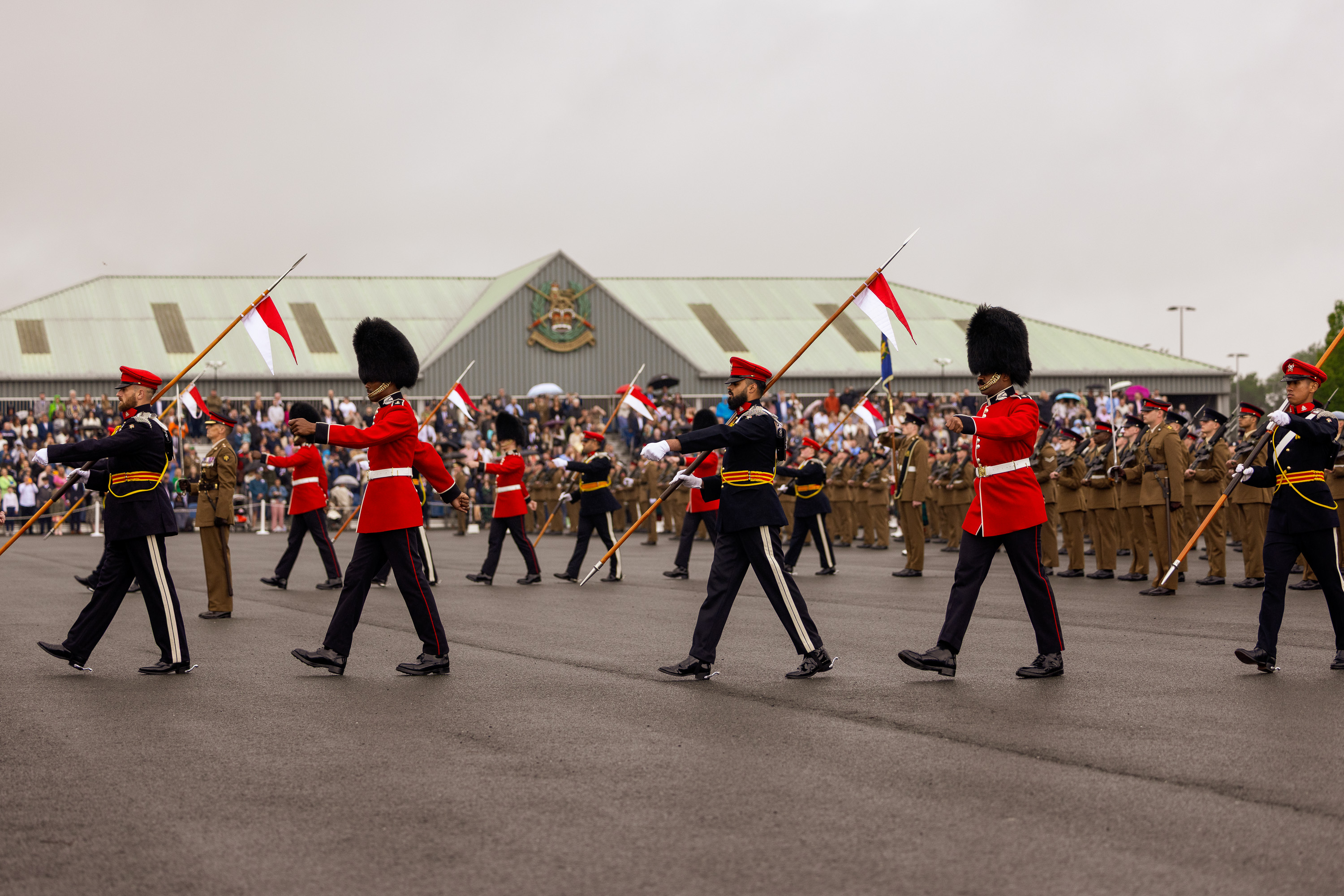 Soldiers in ceremonial dress are marching across the parade square with white and red flags. The graduating soldiers in their brown unifrom are stood in lines behind them. The background shows the AFC Harrogate building with the family and friends crowd in front of the building. 