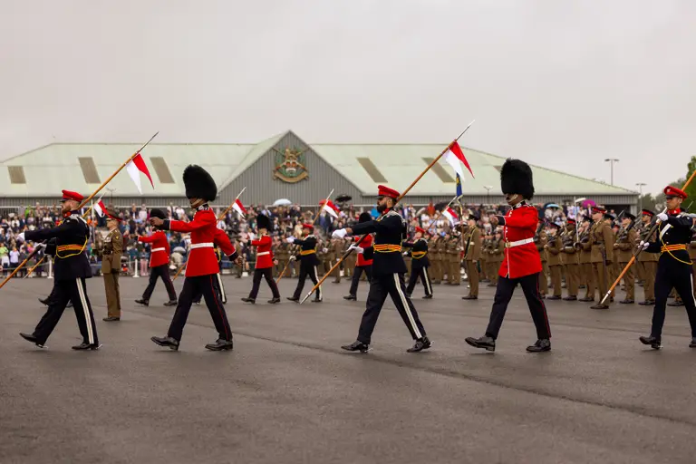 Soldiers in ceremonial dress are marching across the parade square with white and red flags. The graduating soldiers in their brown unifrom are stood in lines behind them. The background shows the AFC Harrogate building with the family and friends crowd in front of the building.