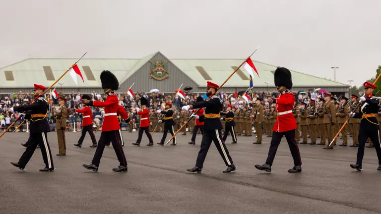 Soldiers in ceremonial dress are marching across the parade square with white and red flags. The graduating soldiers in their brown unifrom are stood in lines behind them. The background shows the AFC Harrogate building with the family and friends crowd in front of the building.