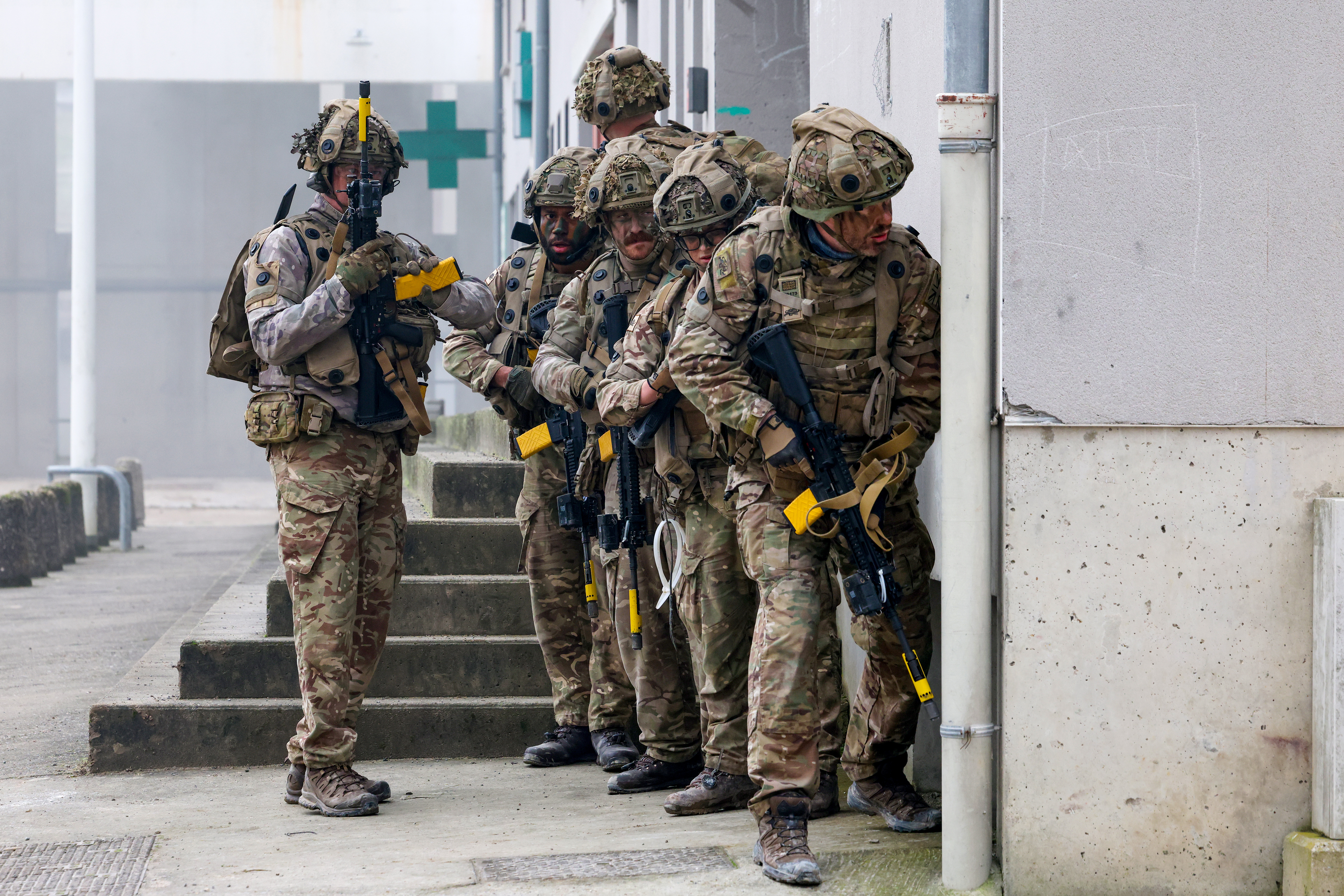 A group of soldiers in camouflage gear standing in formation near a building, holding rifles with yellow markers.