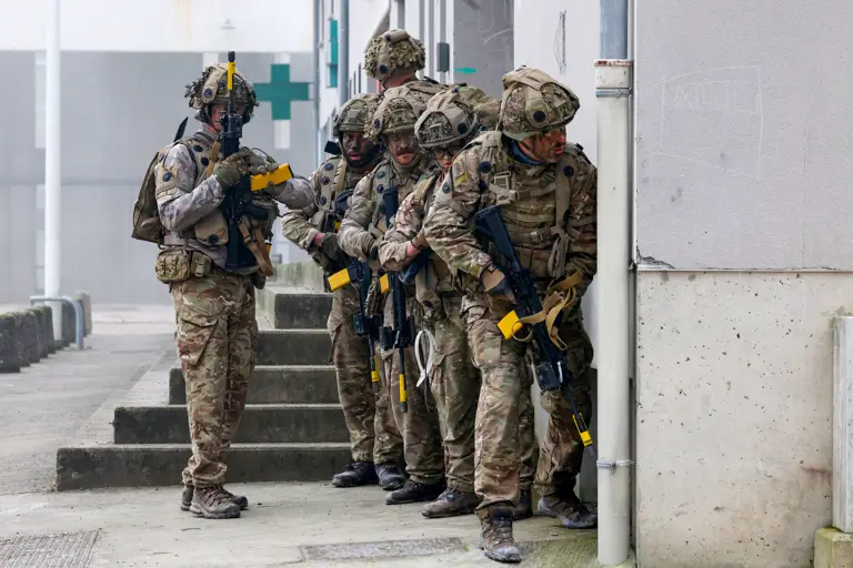 A group of soldiers in camouflage gear standing in formation near a building, holding rifles with yellow markers.